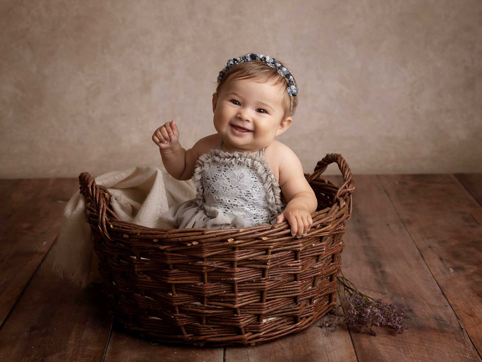 Baby ler stort och tittar in i kameran. Hon sitter i en brun korg och har en blomsterkrans i håret.