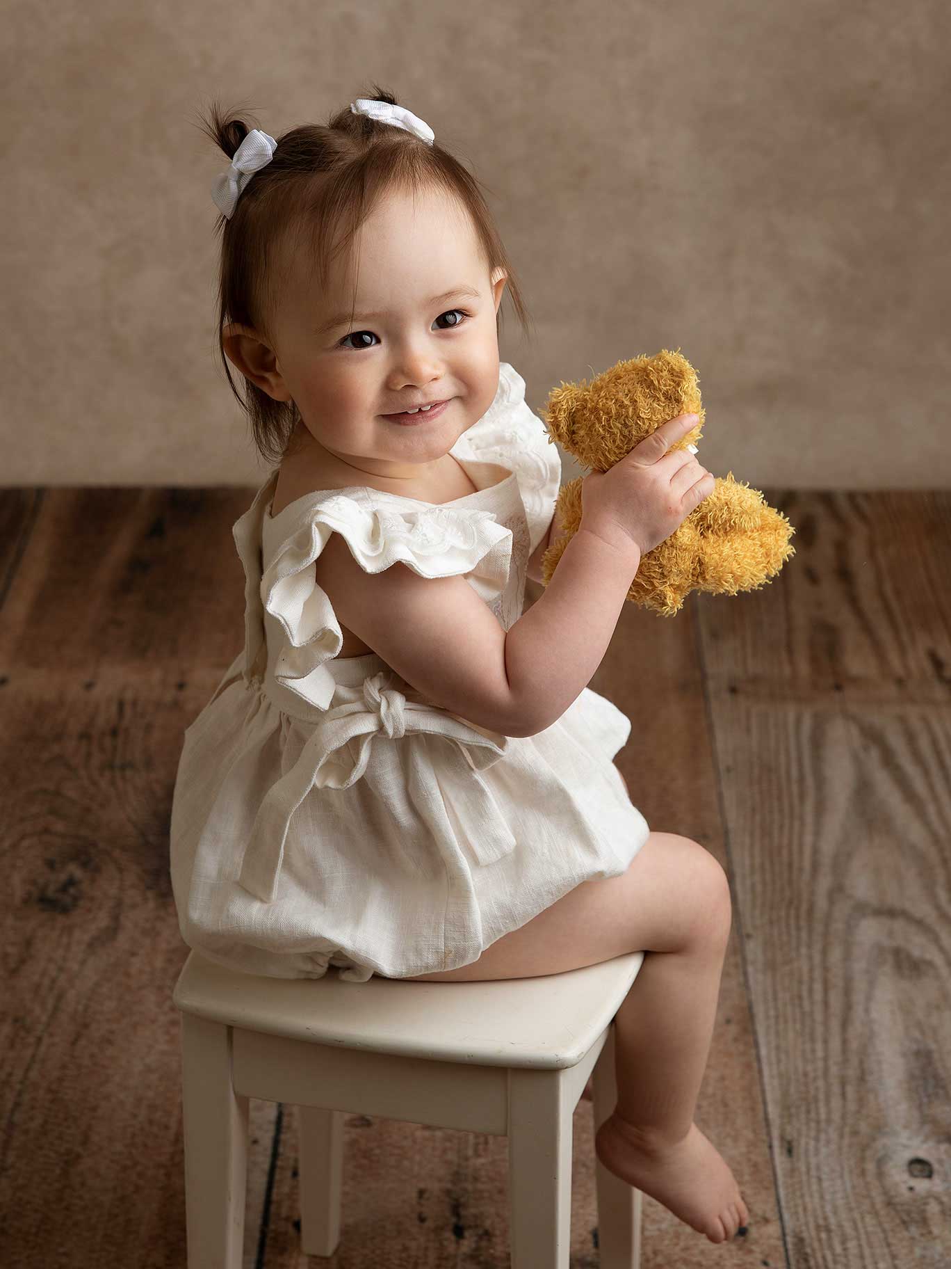 Liten flicka sitter med vita rosetter i håret och håller en nalle i handen. Hon ler och fotografen har fångat henne fint med kameran i studio i Malmö.