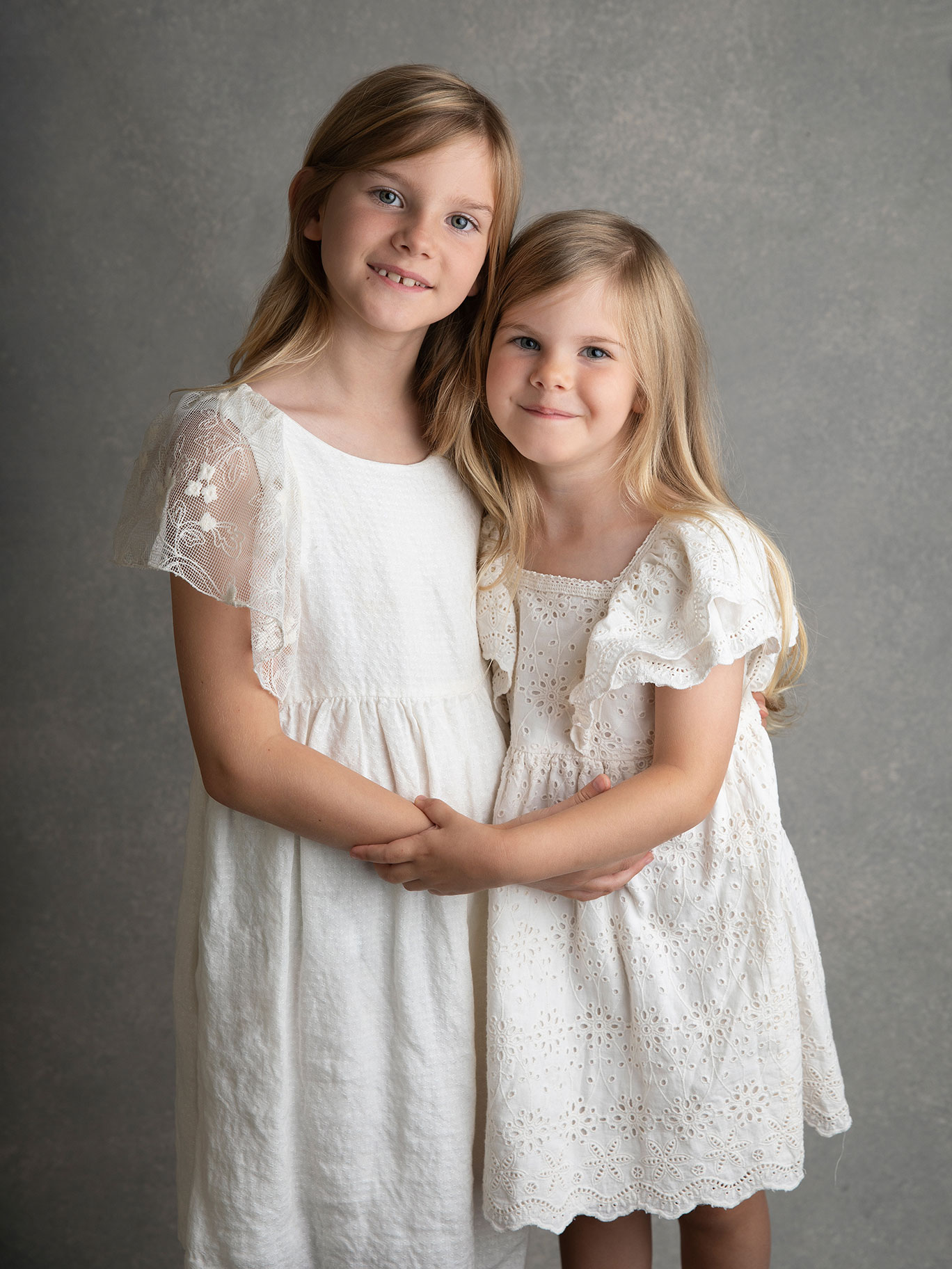 Två systrar i vita klänningar håller hand och blir fotograferade i en studio i Skåne.