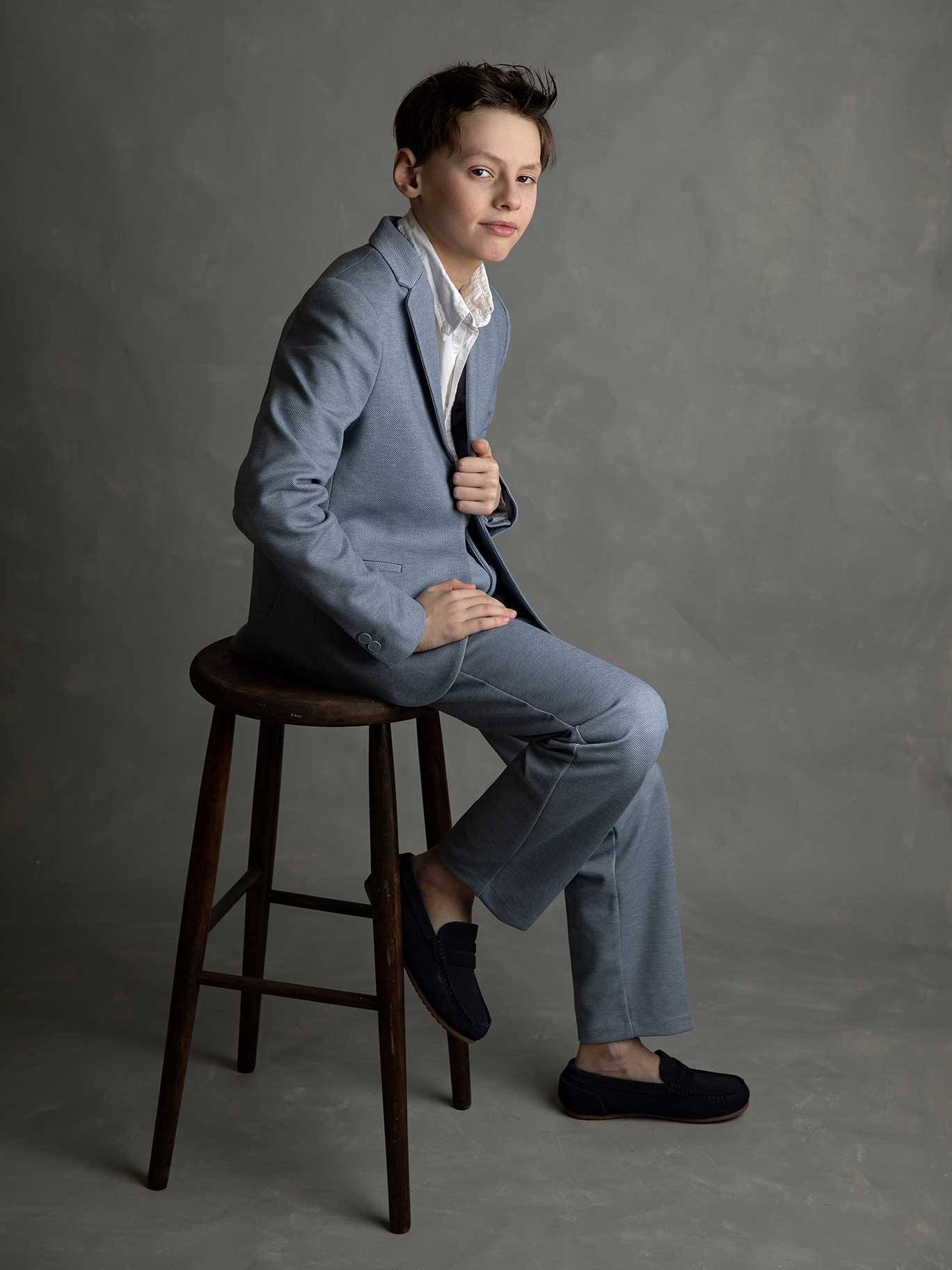 Pojke i blå kostym poserar avslappnat på en pall framför kameran i studio i Malmö.