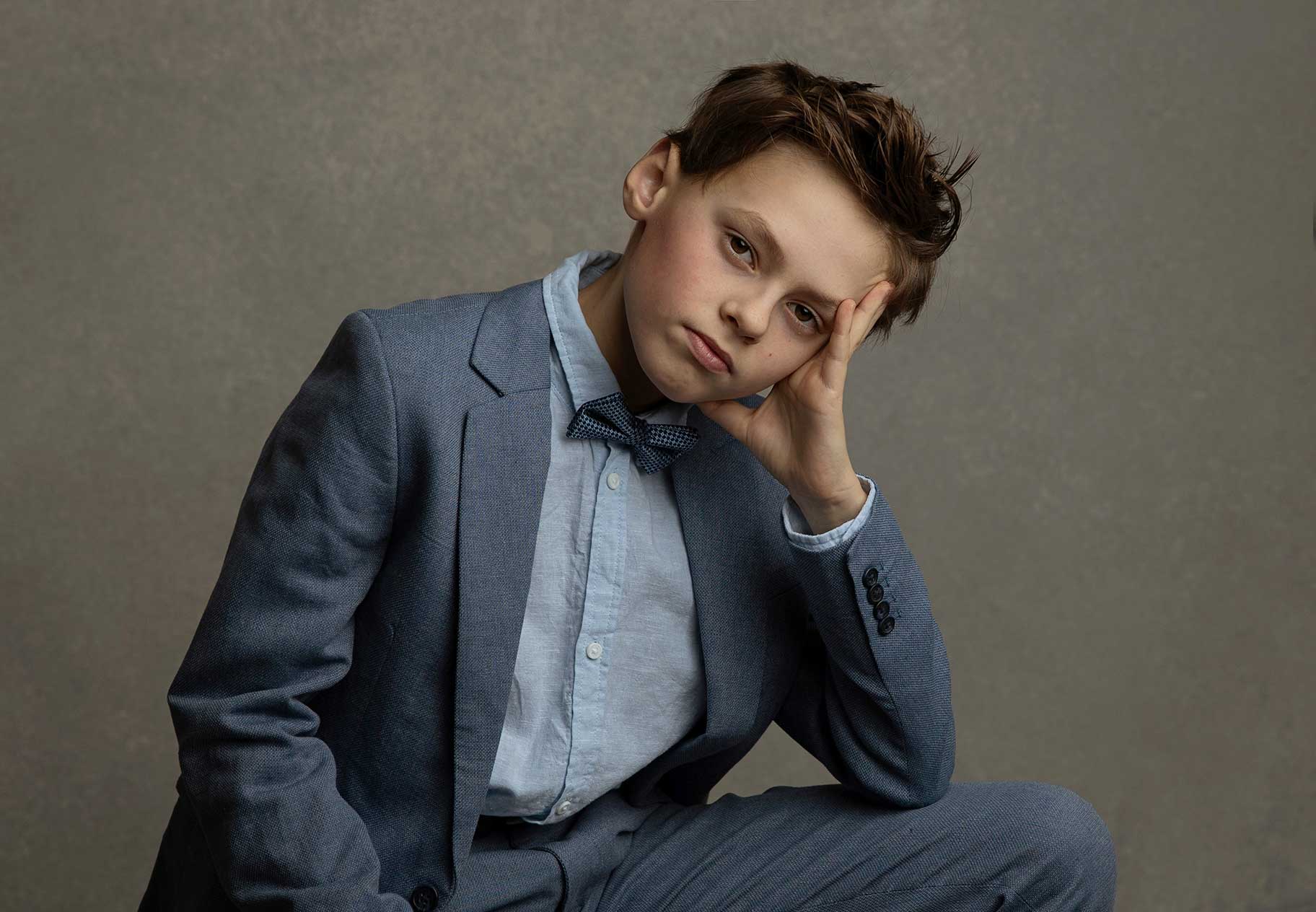 Pojke i blå kostym och fluga poserar avslappnat framför kameran i studio i Malmö.