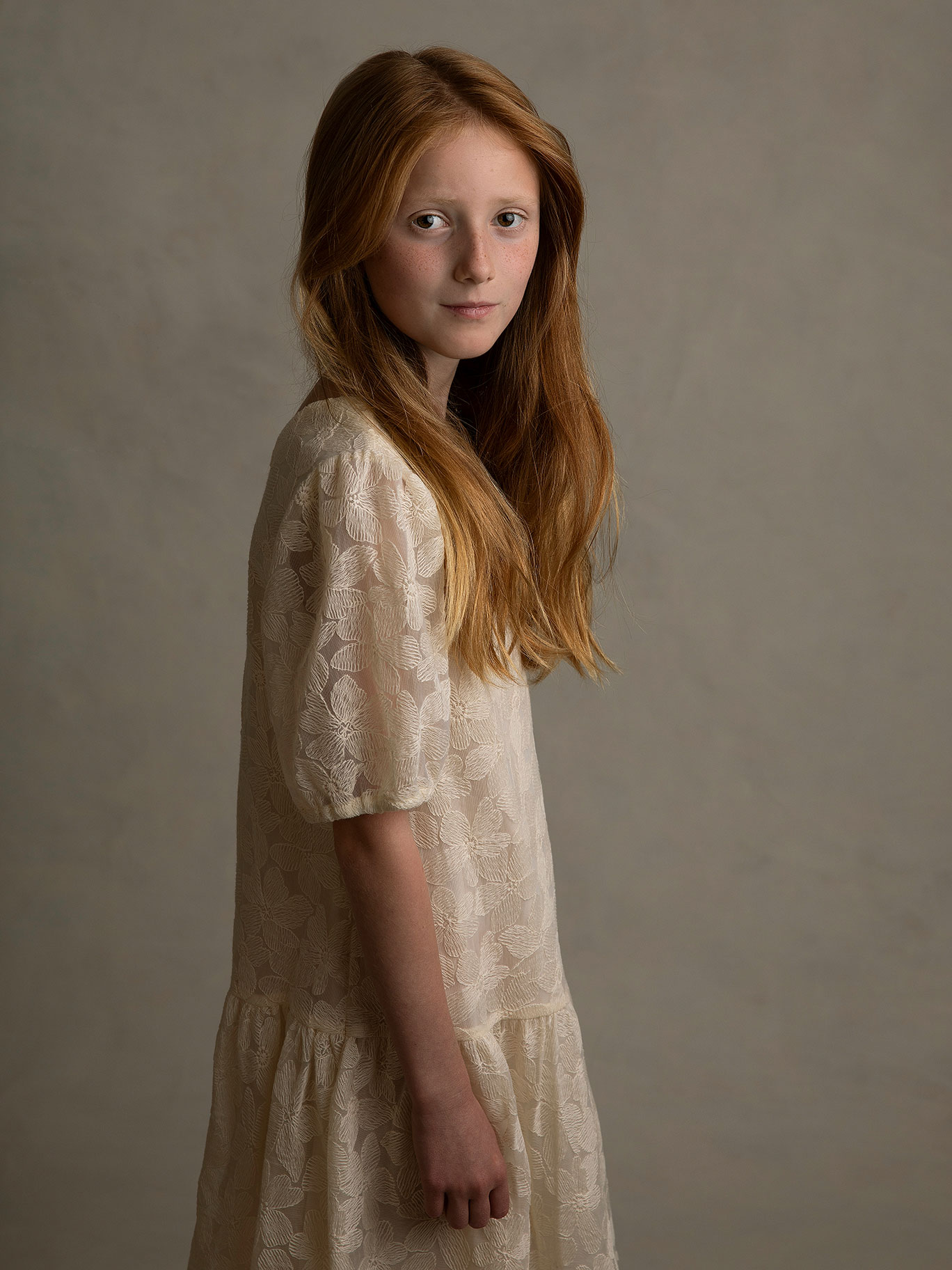 Rödhårig flicka i beige klänning poserar framför ljusgrå bakgrund i Malmö-studio.