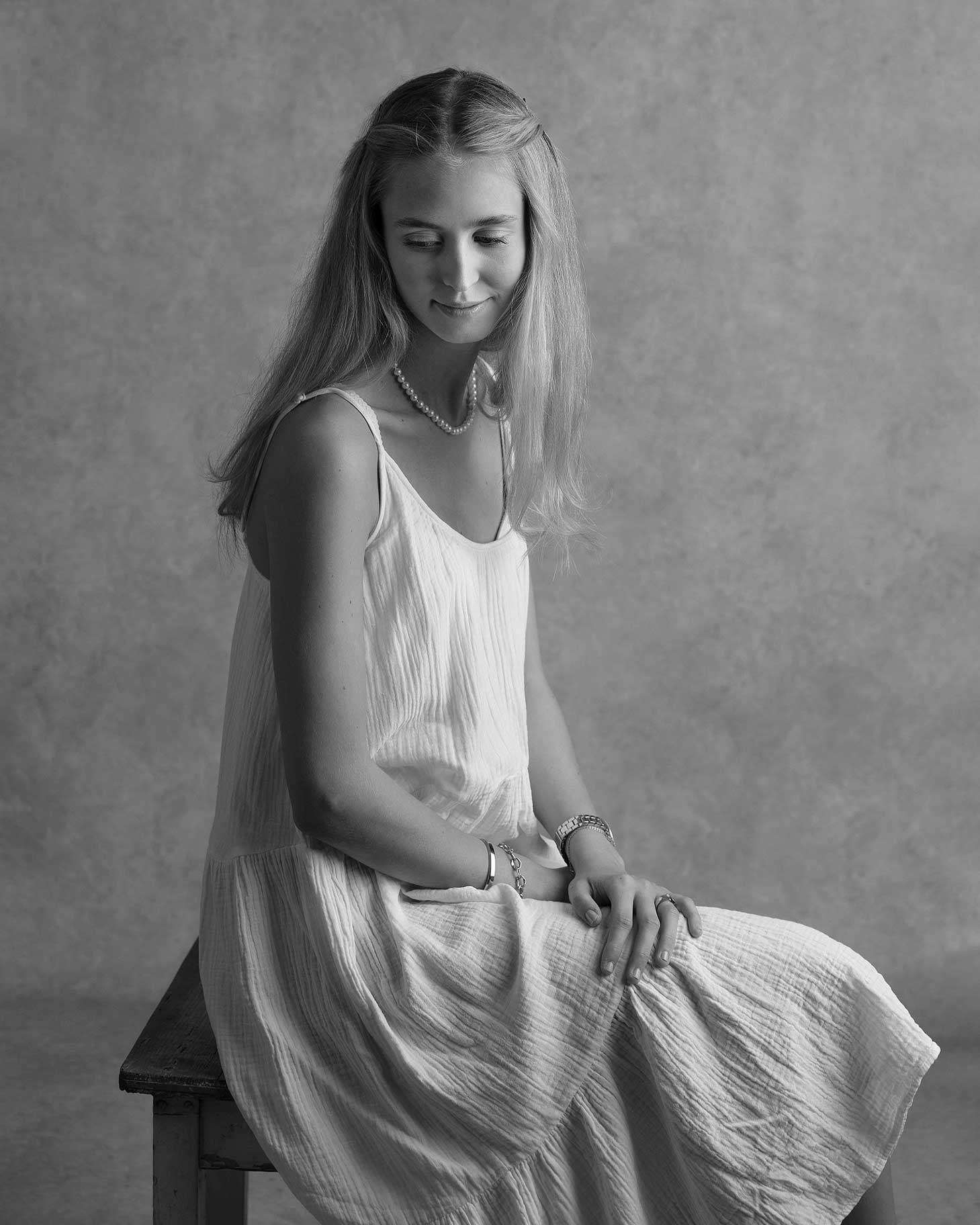 Ung tjej i vit klänning sitter och ler och tittar ner under en fotografering i Malmö.