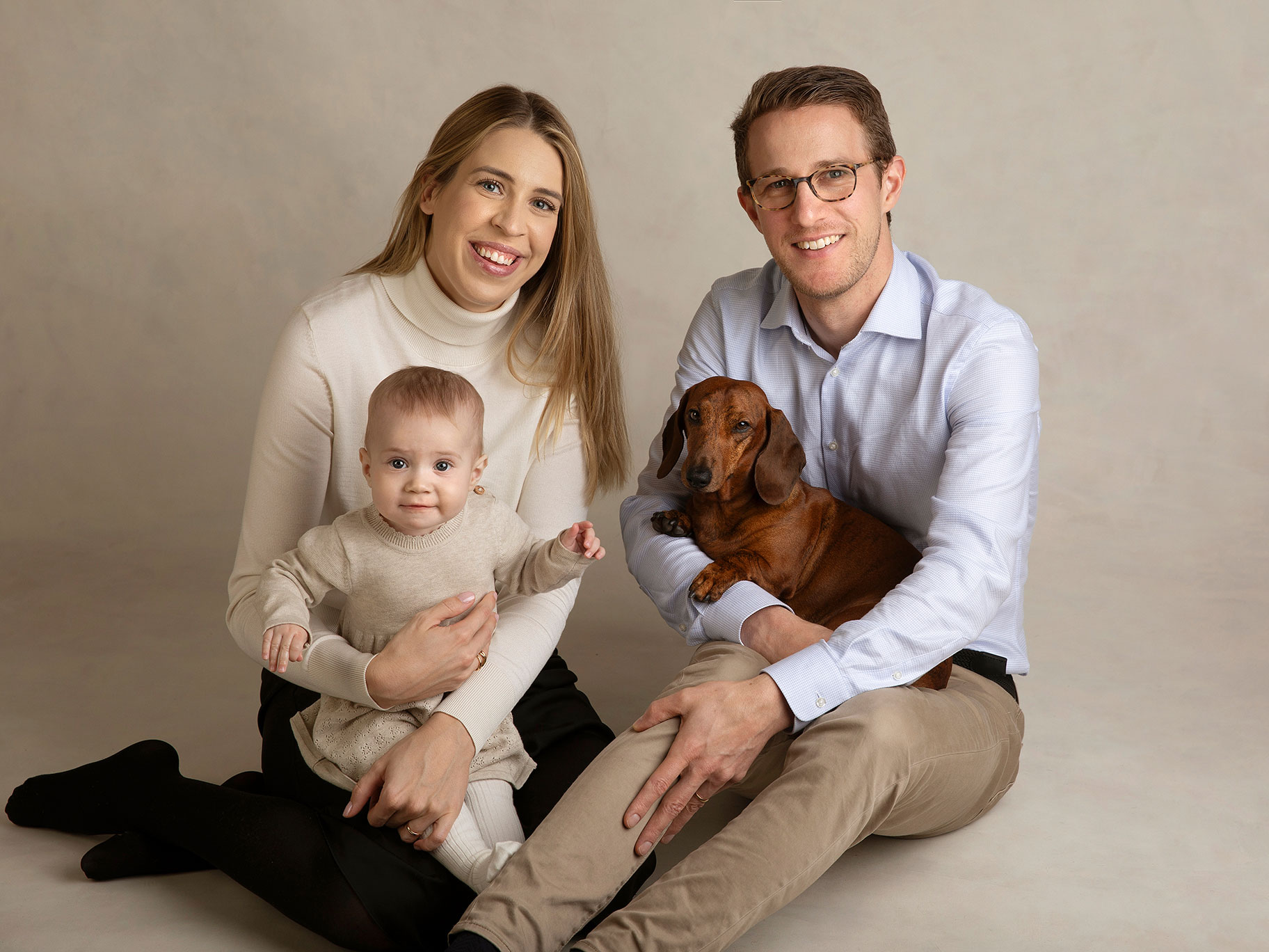 Familj med baby och en hund sitter tätt tillsammans framför grå bakgrund i studion.