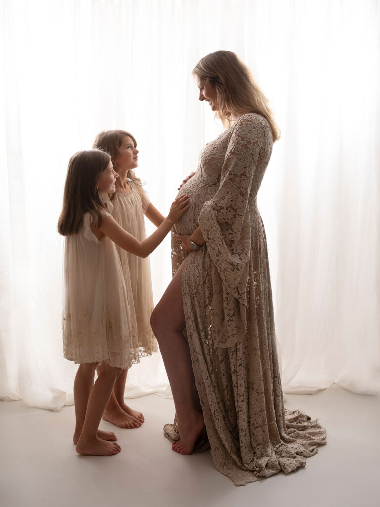 Två flickor håller om sin mammas stora gravidmage och tittar glatt på varandra i studion i Malmö.