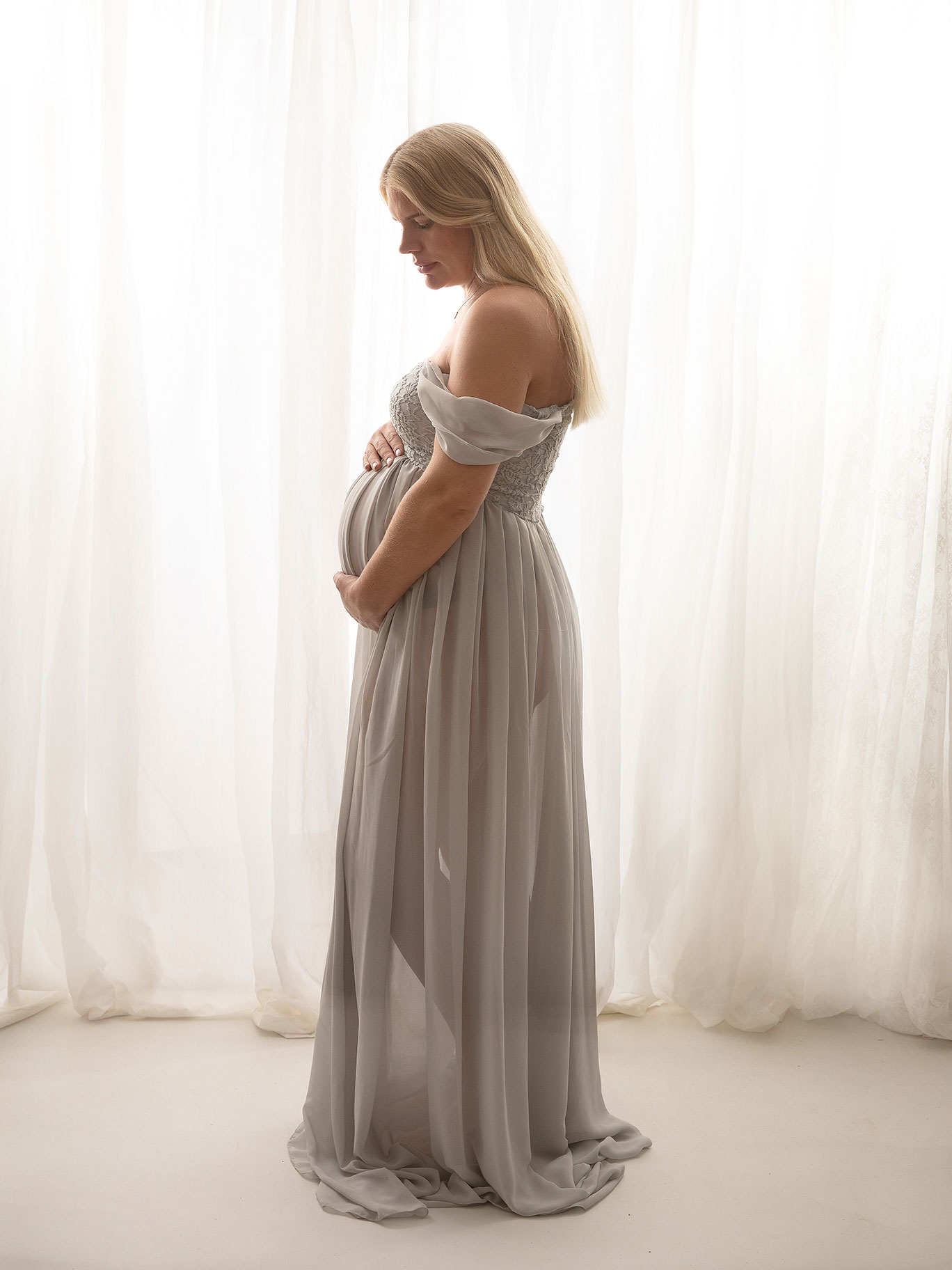 Gravid kvinna i ljusblå klänning poserar framför ljus bakgrund i Fotostudio Anna Arvidsson.