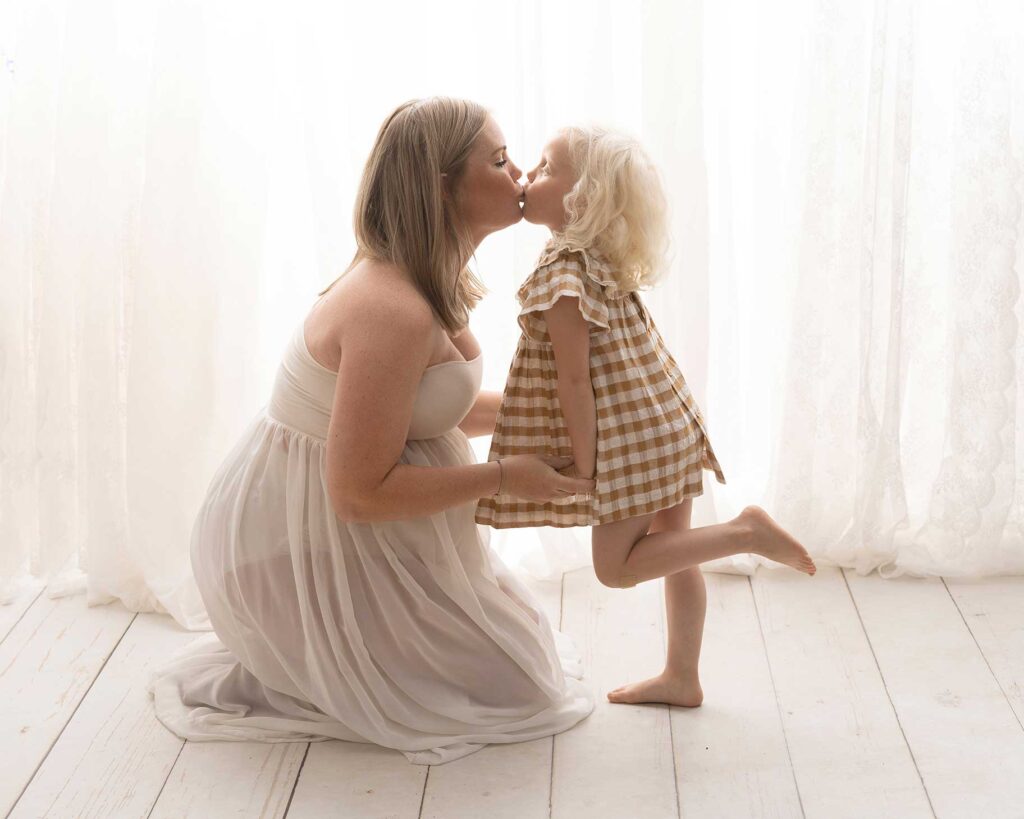 Söt bild på gravid mamma som pussar sin dotter i studion i Malmö med vit bakgrund i motljus.