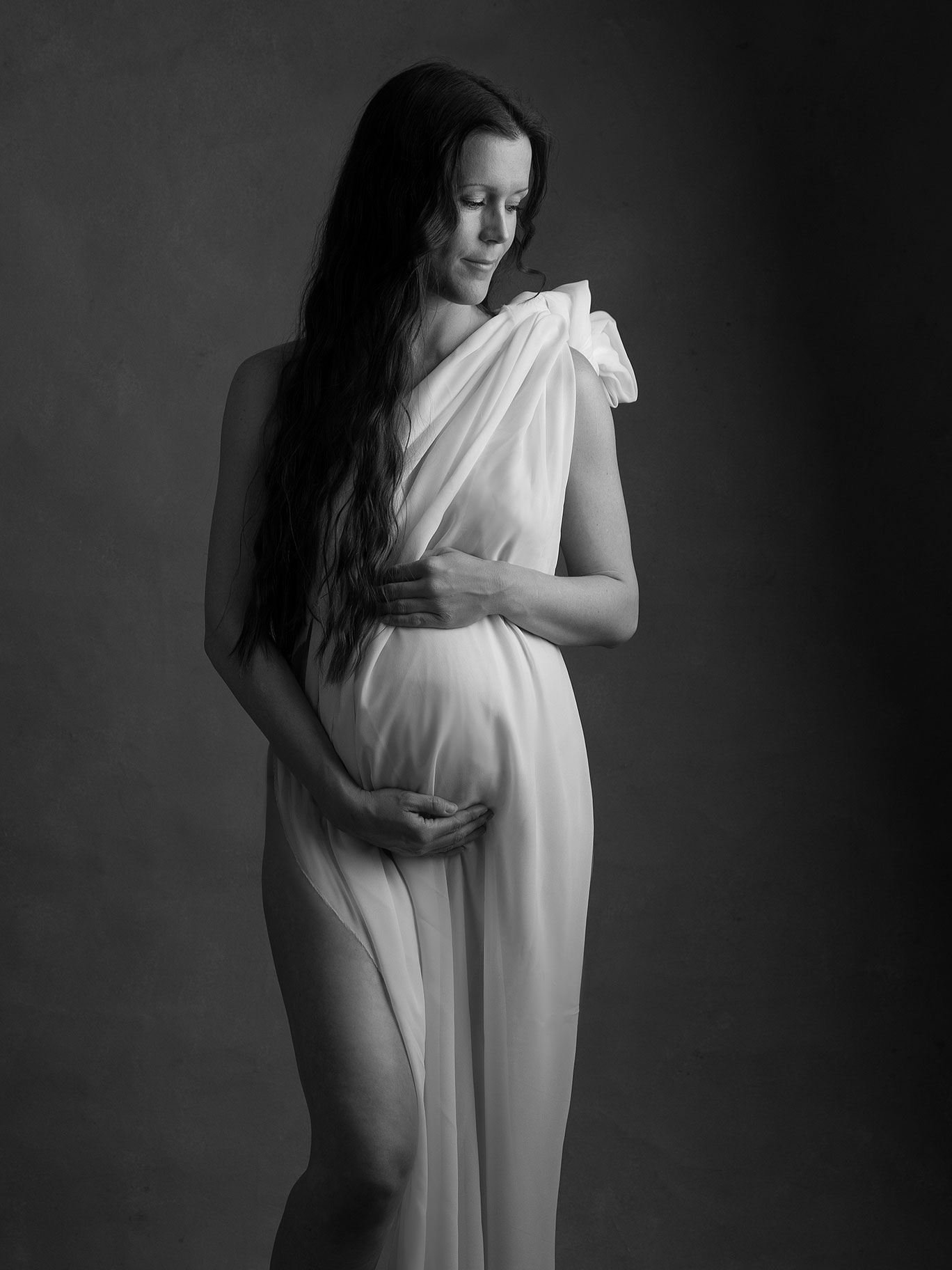 Gravid kvinna med händerna på magen, omgiven av mjukt ljus i studion i Malmö.