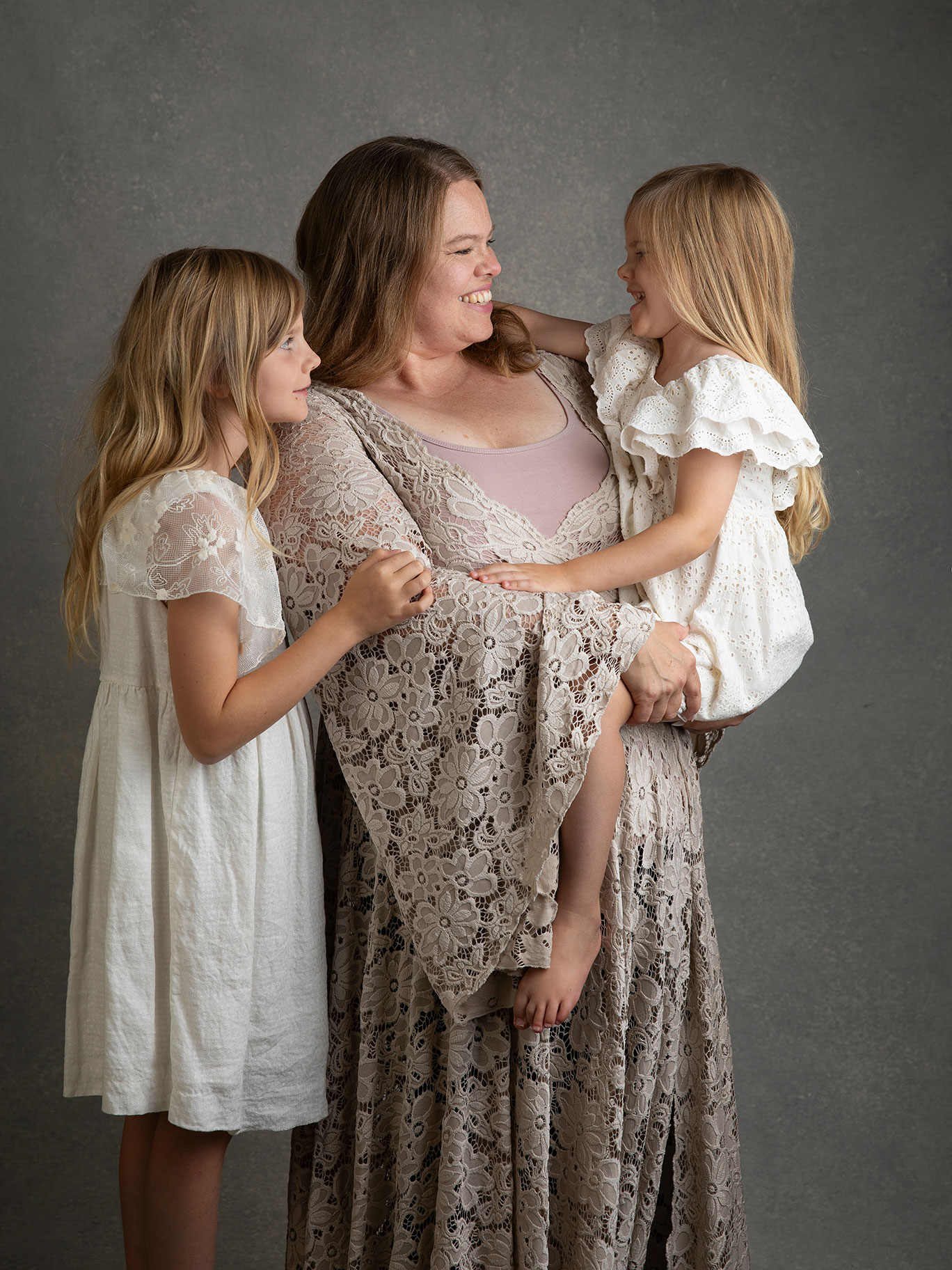 Mamma håller en dotter i famnen och den andra dottern står bredvid och de ser glada ut och busar.