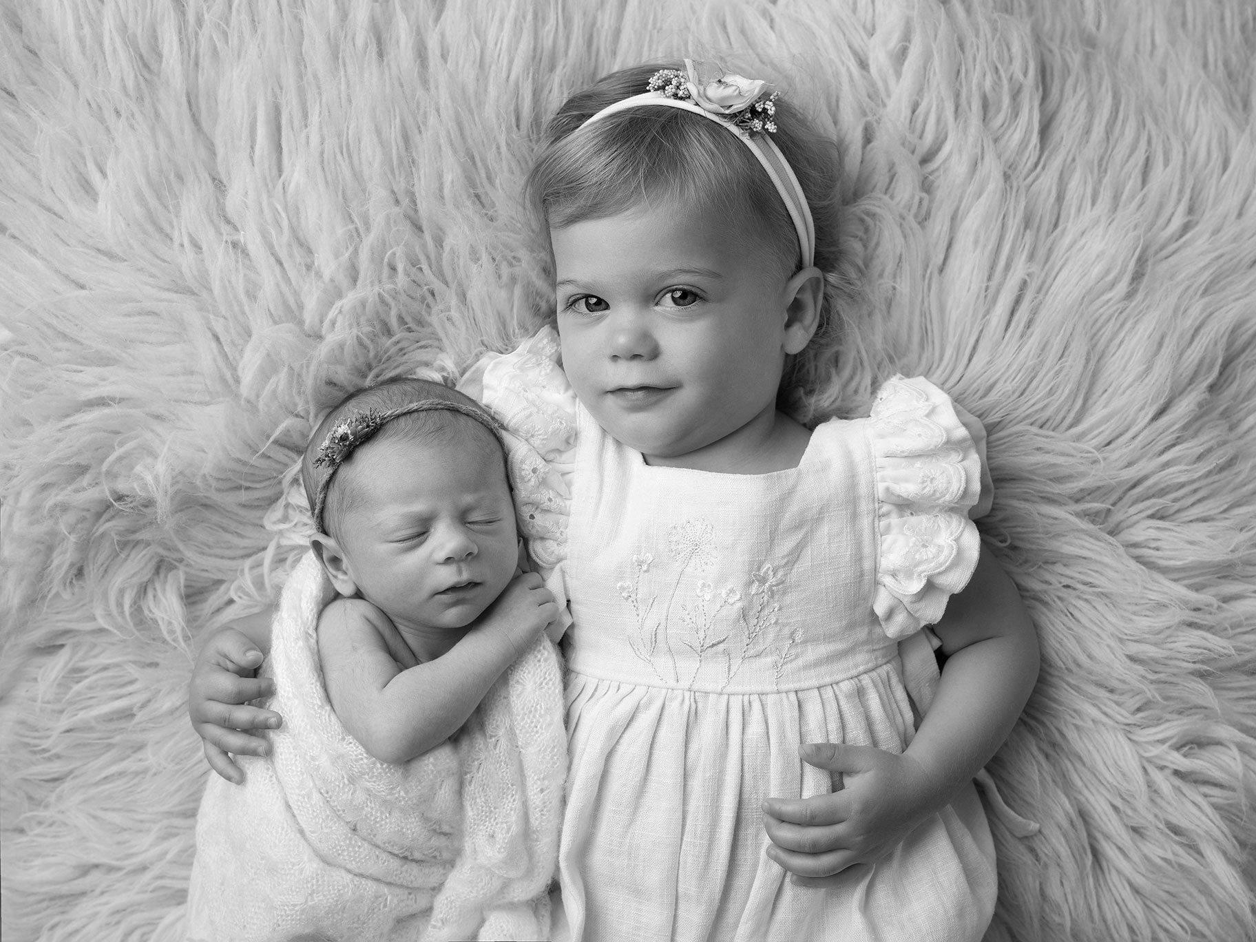 Flicka i söt klänning håller om sin nyfödda lillasyster och tittar in i kameran i studio i Malmö.