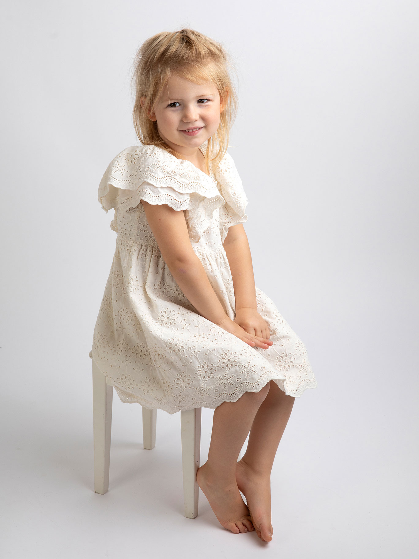 Liten flicka sitter fint på en stol, hon bär en vit klänning och ler.