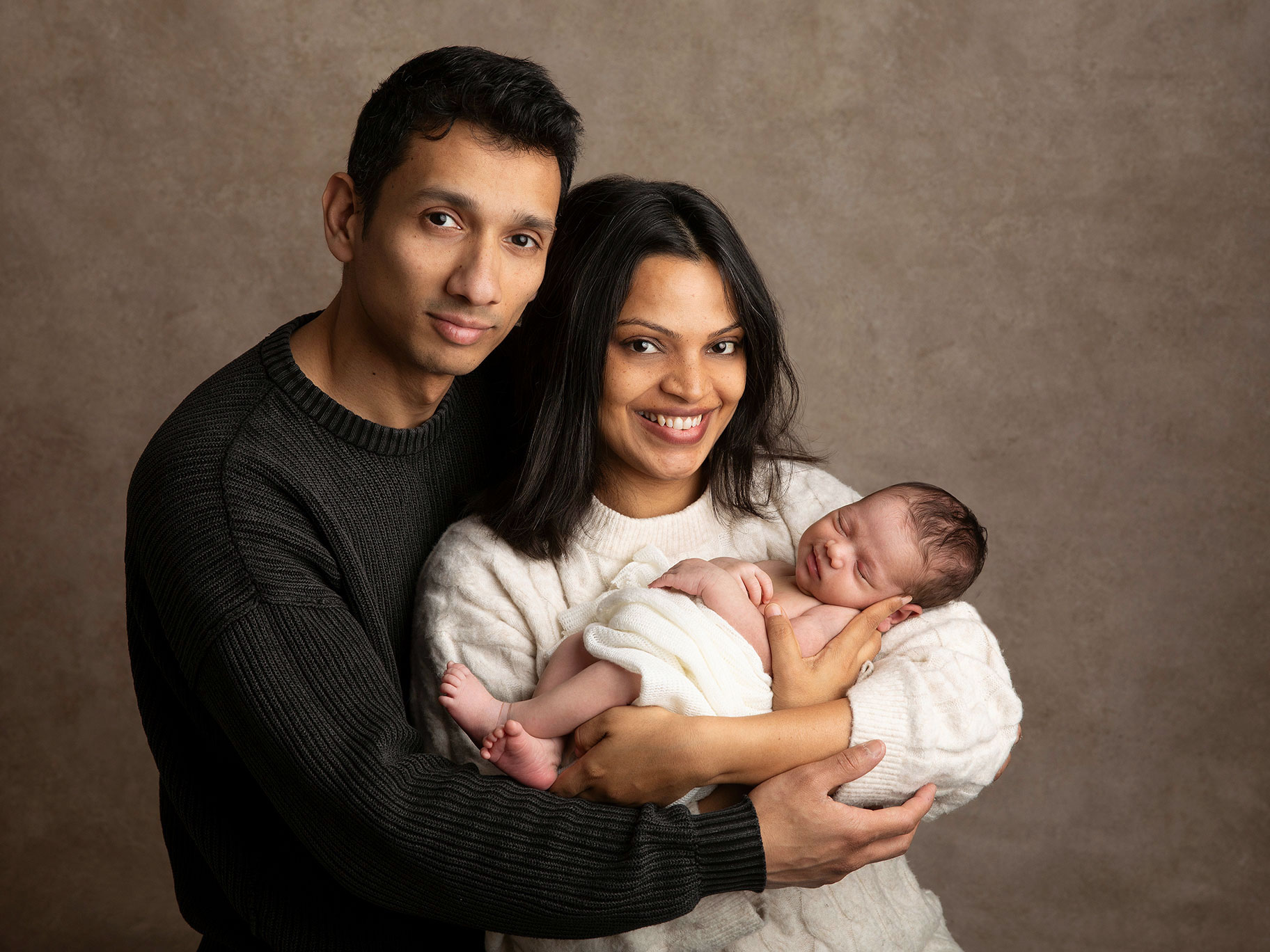 Nyblivna föräldrar tittar stolt in i kameran med sin baby i famnen hos fotografen Anna Arvidsson i Skåne.